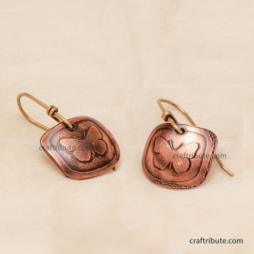 A pair of handmade copper earrings featuring an etched butterfly design on square-shaped dangles, displayed on a textured beige fabric background.