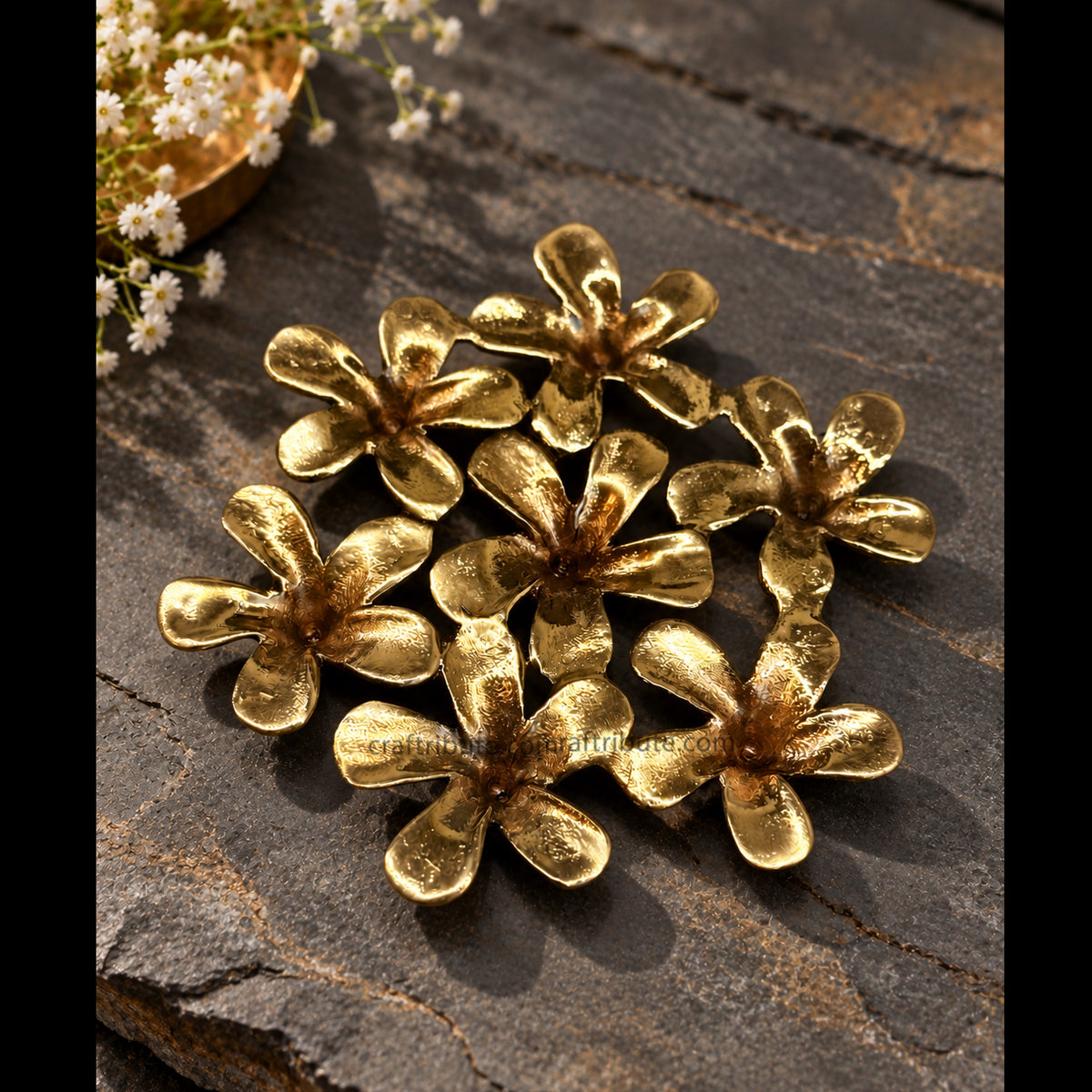 Handcrafted brass Champa flower coaster featuring seven intricately detailed floral elements with an antique gold finish, arranged on a rustic dark stone surface with delicate white flowers.