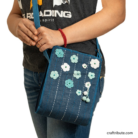 A model carrying a hand embroidered jute sling bag in bue with light blue and white crochet flowers