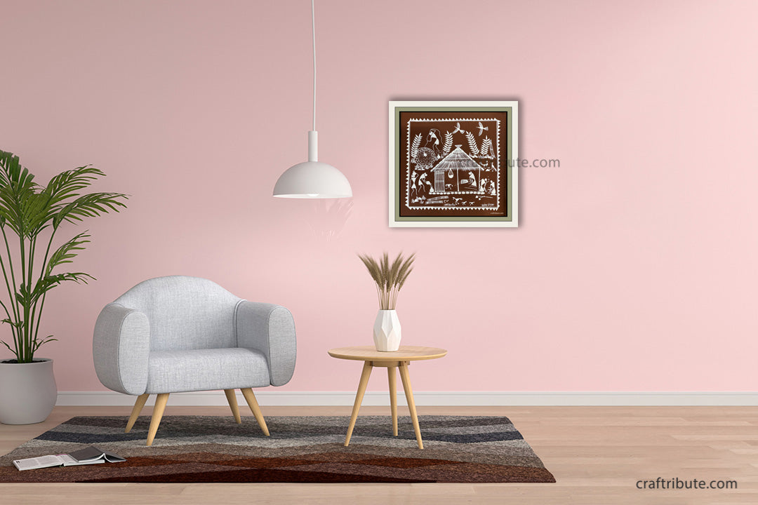 A beautiful framed square Warli painting hangs on a light pink wall in a modern living room mockup with a gray armchair, side table, and hanging lamp.