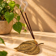 Handcrafted brass betel leaf incense holder with two lit incense sticks releasing delicate wisps of smoke, styled on a warm marble surface with a green heart-shaped leaf plant in the background.