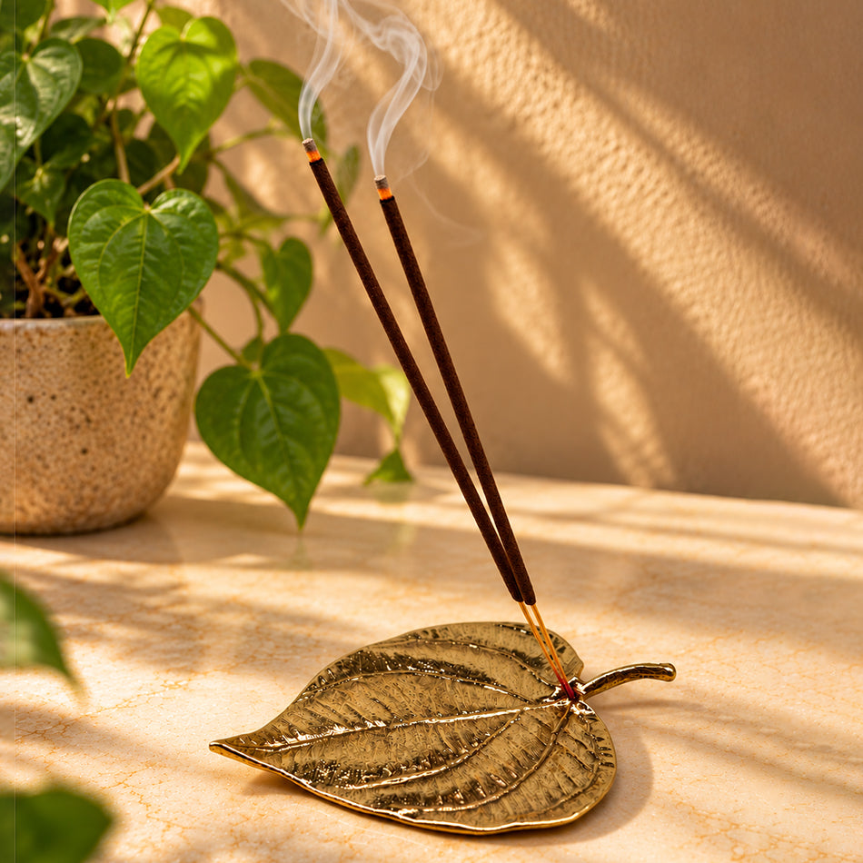 Handcrafted brass betel leaf incense holder with two lit incense sticks releasing delicate wisps of smoke, styled on a warm marble surface with a green heart-shaped leaf plant in the background.