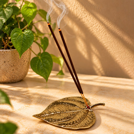 Handcrafted brass betel leaf incense holder with two lit incense sticks releasing delicate wisps of smoke, styled on a warm marble surface with a green heart-shaped leaf plant in the background.