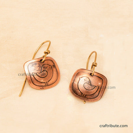 A pair of handmade copper earrings with brass ear wires, featuring an etched bird design on square-shaped dangles, displayed on a textured beige fabric background.