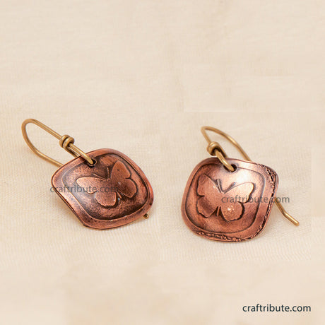 A pair of handmade copper earrings featuring an etched butterfly design on square-shaped dangles, displayed on a textured beige fabric background.