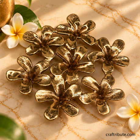 Handcrafted brass Champa flower coaster featuring seven intricately detailed floral elements with an antique gold finish, styled on cream marble with fresh Champa flowers and green leaves.