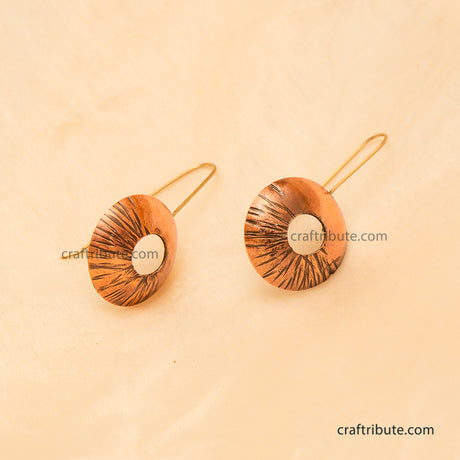 A pair of handmade copper earrings with a textured, sunburst-like pattern displayed on a warm beige background.