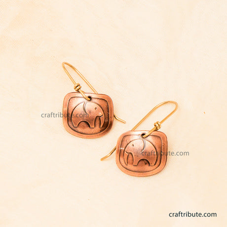A pair of handmade copper earrings with an etched elephant design, displayed on a light beige textured background.