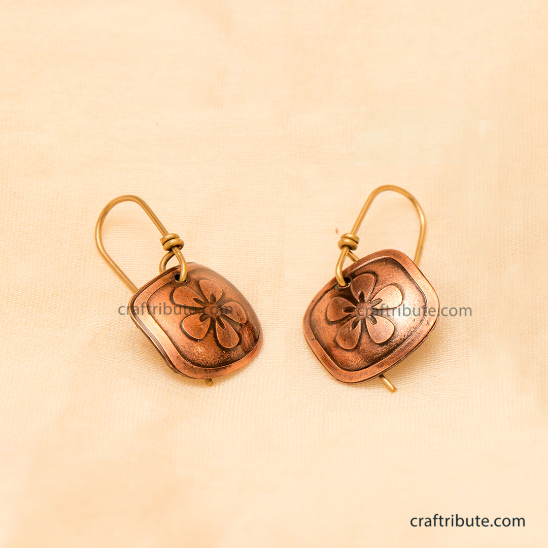 A pair of handmade copper drop earrings with an etched flower design, displayed on a textured beige fabric background.