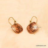 A pair of handmade copper drop earrings with an etched flower design, displayed on a textured beige fabric background.