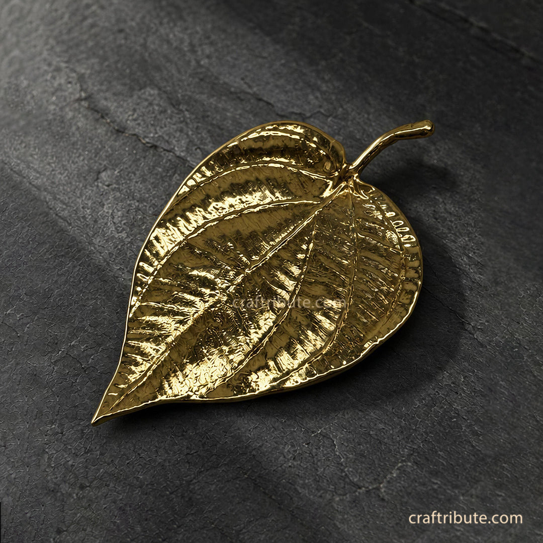 Handcrafted brass betel leaf décor with intricate vein detailing and an antique gold finish, displayed on a textured black stone surface.