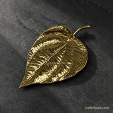 Handcrafted brass betel leaf décor with intricate vein detailing and an antique gold finish, displayed on a textured black stone surface.