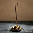 Handcrafted brass lotus incense holder with two incense sticks, featuring intricately detailed petals and an antique gold finish, displayed against a rustic stone backdrop.