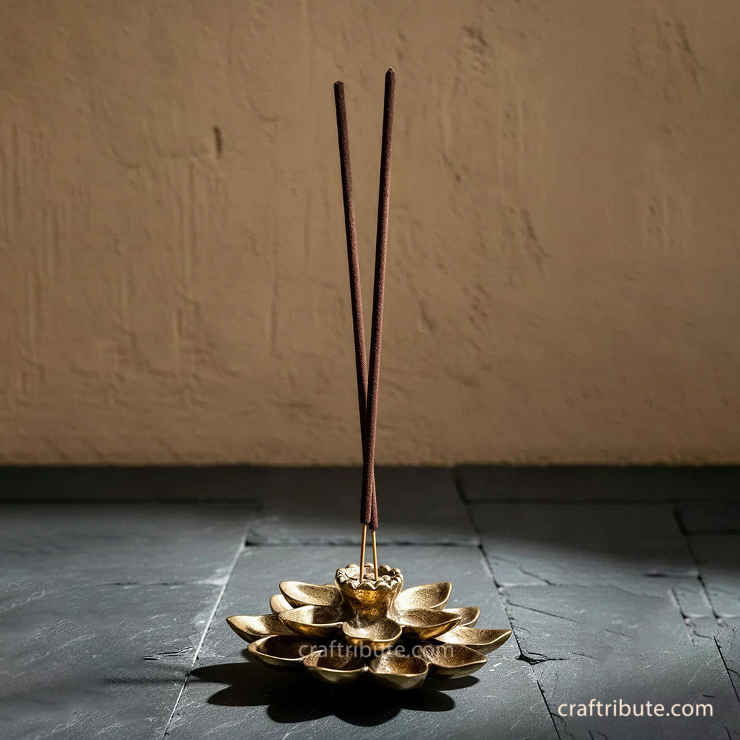 Handcrafted brass lotus incense holder with two incense sticks, featuring intricately detailed petals and an antique gold finish, displayed against a rustic stone backdrop.
