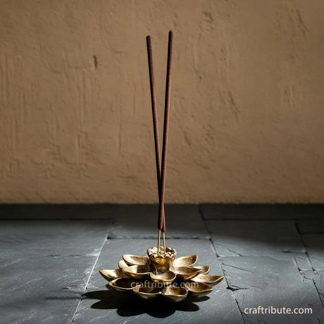 Handcrafted brass lotus incense holder with two incense sticks, featuring intricately detailed petals and an antique gold finish, displayed against a rustic stone backdrop.